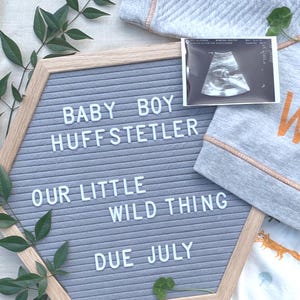 以下が含まれることがあります： 白い文字で「BABY BOY HUFFSTETLER, OUR LITTLE WILD THING, DUE JULY」と書かれた灰色のフェルトレターボード。超音波画像とベビー服も画像にあります。ボードは木製のフレームと緑に囲まれています。