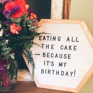 May include: A white felt letter board in a wooden frame with the text "EATING ALL THE CAKE BECAUSE IT'S MY BIRTHDAY!". A bouquet of flowers in a glass vase is to the left of the board.