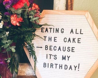 May include: A white felt letter board in a wooden frame with the text "EATING ALL THE CAKE BECAUSE IT'S MY BIRTHDAY!". A bouquet of flowers in a glass vase is to the left of the board.