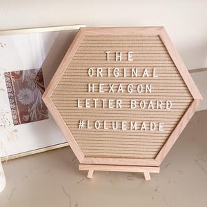 May include: A light brown hexagon letter board with a wooden frame and stand. The board features the text "THE ORIGINAL HEXAGON LETTER BOARD #LOLUEMADE" in white letters. A framed picture is in the background.