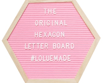 May include: A pink hexagon letter board with a light wood frame. The board has horizontal grooves and white plastic letters spelling "THE ORIGINAL HEXAGON LETTER BOARD #LOLUEMADE".