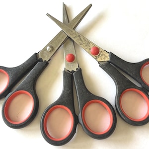 May include: Close-up of five scissors with black handles, red accents, and stainless steel blades. The scissors are arranged in a fan shape against a white background. The blades are crossed, and the handles have finger holes.