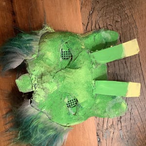 May include: A handmade green mask with a textured surface, featuring eye openings with a grid pattern. The mask has green and blue faux fur accents and green painted extensions with yellow tips. The mask is likely for costume or cosplay.