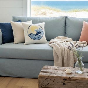 May include: A light blue sofa featuring a collection of throw pillows in navy, cream, and peach, including one with a circular embroidered wave and sun design. A beige knit throw blanket is draped over the seat, and a glass vase sits nearby.