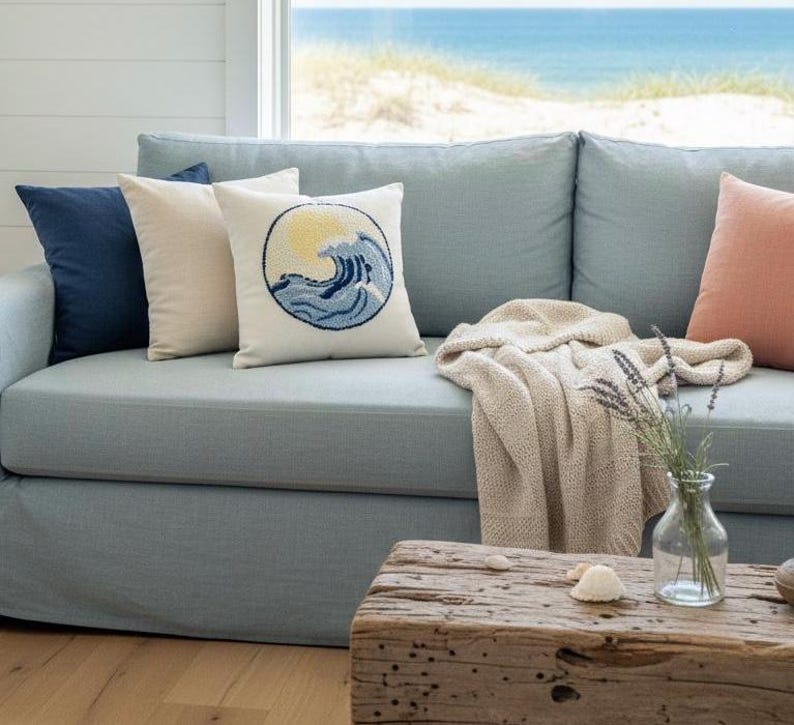 May include: A light blue sofa featuring a collection of throw pillows in navy, cream, and peach, including one with a circular embroidered wave and sun design. A beige knit throw blanket is draped over the seat, and a glass vase sits nearby.