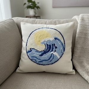 May include: A square off-white throw pillow featuring a circular punch needle embroidery design. The design depicts a stylized ocean wave in shades of light and dark blue, accented by a pale yellow sun against a textured, cream-colored background.