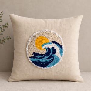 May include: A square beige throw pillow featuring a circular punch needle embroidery design in the center. The design depicts a bright yellow sun positioned above a stylized ocean wave rendered in shades of dark blue, light blue, and white.