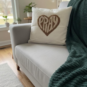 May include: A cream cushion featuring a brown heart design with the word "PRAY" in the centre. It rests on a light grey sofa, with a dark green blanket draped over it. The room has a neutral colour palette.