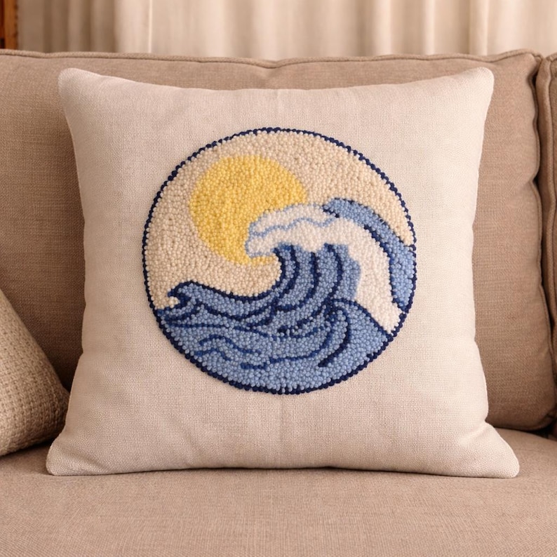 May include: A square throw pillow with a beige fabric cover featuring a circular punch needle embroidery design. The design depicts a yellow sun positioned behind a large, textured blue and white ocean wave, all outlined in a dark blue thread.