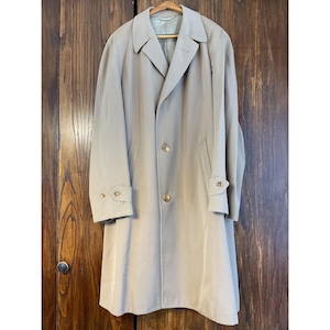 Peut inclure: Un trench-coat beige au design classique, avec un col cranté, des boutons et des poignets réglables. Le manteau est suspendu à un cintre en bois devant une porte en bois. Le manteau a un ourlet à la hauteur du genou.