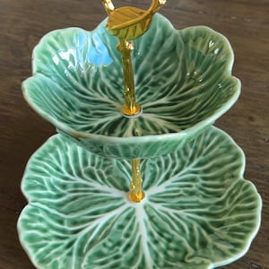 May include: Two-tiered serving dish with a leaf design. The green ceramic dishes are supported by a gold-coloured metal stand and handle. The leaf pattern is detailed with visible veins.
