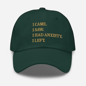 Puede incluir: Gorra de béisbol verde oscuro con el texto dorado: "I CAME. I SAW. I HAD ANXIETY. I LEFT." La gorra tiene una visera curva y un botón en la parte superior. Un accesorio casual.
