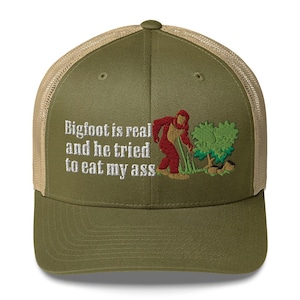 Può includere: Cappellino da camionista verde oliva con retro in rete beige. Il davanti presenta la scritta ricamata "Bigfoot is real and he tried to eat my ass" con un'immagine di Bigfoot.