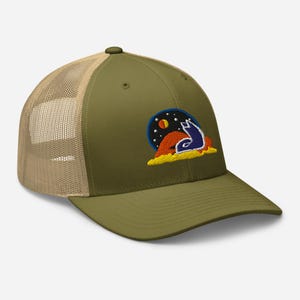 May include: Olive green trucker hat with a tan mesh back and a curved brim. The hat features an embroidered patch with a blue wolf howling at a night sky with a red sun and yellow clouds. The hat has a snapback closure.