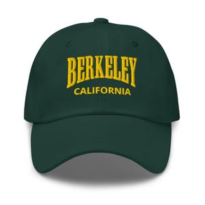 May include: A dark green baseball cap featuring the gold embroidered word "BERKELEY" arched across the front, with "CALIFORNIA" below. The cap has a curved peak and a structured crown.