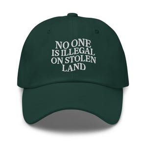 May include: A dark green baseball cap featuring the embroidered phrase "NO ONE IS ILLEGAL ON STOLEN LAND" in white. The cap has a curved peak and an adjustable strap at the back.