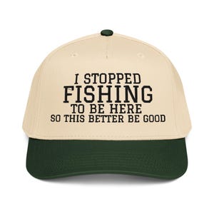 May include: A beige and dark green baseball cap with the text "I STOPPED FISHING TO BE HERE SO THIS BETTER BE GOOD" in black. The cap has a curved brim and a button on top.