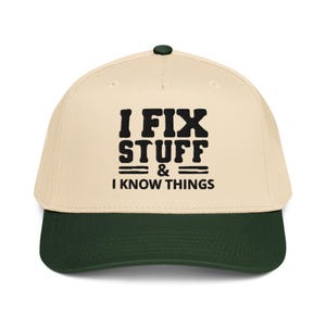 May include: A beige baseball cap with a dark green brim and button. The cap features the text "I FIX STUFF & I KNOW THINGS" in bold black letters. The hat is a classic style, suitable for casual wear.