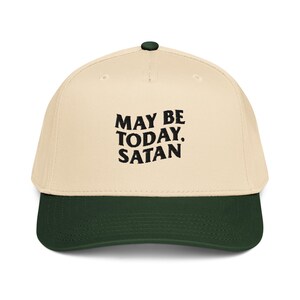 May include: A baseball cap with a cream-colored crown and a dark green brim. The cap features the text "MAY BE TODAY. SATAN" in black, bold letters. The cap has a dark green button on top.
