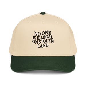 Può includere: Un cappellino da baseball con corona color crema e visiera verde scuro. Il cappellino presenta il testo ricamato "NO ONE IS ILLEGAL ON STOLEN LAND" in nero. Il cappellino ha un bottone verde scuro sulla parte superiore.