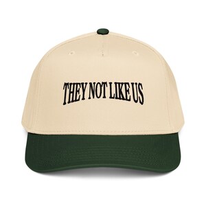May include: A baseball cap with a cream-colored crown and a dark green brim. The cap features the embroidered text "THEY NOT LIKE US" in black, arched across the front. The cap has a dark green button on top.