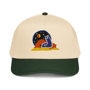 May include: Cream-colored baseball cap with a dark green brim and button. The cap features an embroidered design of a blue wolf howling at a moon and stars over an orange landscape.