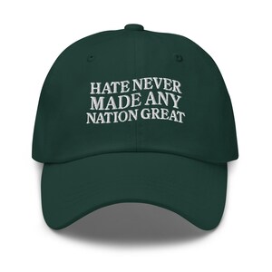 以下が含まれることがあります： 白い刺繍で「HATE NEVER MADE ANY NATION GREAT」と書かれた、濃い緑色の野球帽。帽子にはカーブしたつばと、上部にボタンが付いています。カジュアルなアクセサリーです。