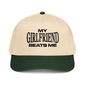 May include: A baseball cap with a cream-colored crown and a dark green brim. The cap features the text "MY GIRLFRIEND BEATS ME" in bold, black letters across the front. The cap has a dark green button on top.