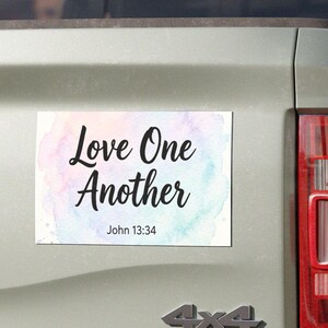 Puede incluir: Una pegatina rectangular con el texto "Love One Another" en escritura negra sobre un fondo de acuarela de colores pastel. El texto "John 13:34" está debajo. La pegatina está adherida a la parte trasera de una camioneta verde claro.