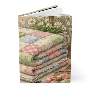 May include: A hardcover journal featuring a watercolor illustration of a stack of folded quilts in pastel colors. The journal's cover also depicts a vase of daisies. A spool of thread rests on the wooden surface.