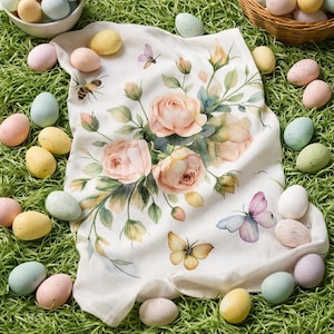 Peut inclure: Un torchon de cuisine blanc à motifs floraux avec un motif aquarelle de roses roses, de feuilles vertes et de papillons. Le torchon est entouré d'œufs de Pâques aux couleurs pastel sur de l'herbe verte.