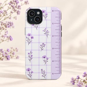 May include: A white phone case featuring a light purple grid pattern and small purple floral illustrations. The right side of the case displays a vertical ruler graphic with markings from 0 to 5 inches, printed in black on a light purple strip.