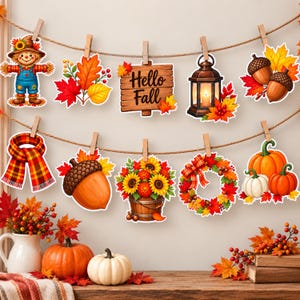 Può includere: Un set di ritagli di carta a tema autunnale appesi a due spaghi con mollette di legno. I ritagli raffigurano uno spaventapasseri, foglie autunnali, un cartello con la scritta Hello Fall, una lanterna, ghiande, una sciarpa a quadri, girasoli, una ghirlanda e zucche.