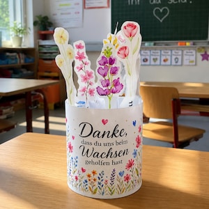 May include: A cylindrical white container decorated with colorful floral illustrations and the German text "Danke, dass du uns beim Wachsen geholfen hast." Several paper cutouts of various flowers in shades of pink, yellow, and purple are placed inside.