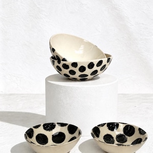 Pode incluir: Três tigelas de cerâmica com uma base de cor creme e bolinhas pretas pintadas à mão. Uma tigela está empilhada sobre a outra, enquanto duas tigelas adicionais estão colocadas lado a lado na frente. Design liso e arredondado.
