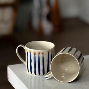 May include: Two ceramic mugs with a cream-colored base and blue vertical stripe design. One mug is upright, the other is on its side. The mugs have a simple handle and a brown accent at the top. The mugs are on a white surface.