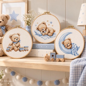 May include: Three circular wooden embroidery hoops containing cross-stitch patterns of teddy bears. One bear sits with a toy train, another sleeps on a blue pillow, and the third rests on a crescent moon. The text reads UKDesign FR.