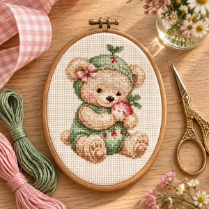 Puede incluir: Un bastidor de bordado ovalado de madera que contiene un patrón de punto de cruz de un oso de peluche marrón claro con un gorro verde y un conjunto a juego. El oso sostiene una pequeña flor rosa sobre tela blanca con cuadrícula.