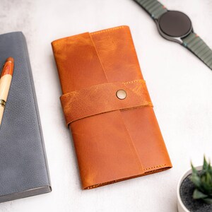 May include: A close-up shot of a brown leather journal with a strap closure and a button. A gray notebook and a pen with a wooden barrel are to the left. A smartwatch and a small potted plant are in the background. The journal is the focal point.