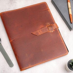 May include: A brown leather portfolio with a strap closure. The portfolio has a distressed finish and visible stitching around the edges. A gray watch with a black face and a small succulent plant in a white pot are also visible.