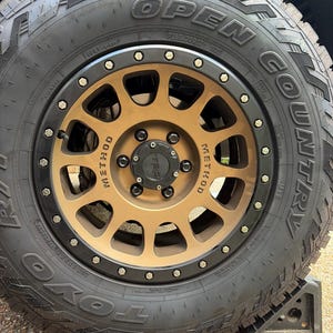 May include: A bronze-colored Method wheel with a black outer ring and silver bolts, fitted with a black Toyo Open Country tire. The center cap features the TRD logo in black, and the tire sidewall displays the text P285/70R17 117S.