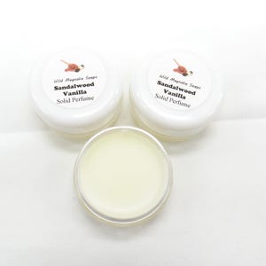 Op de afbeelding: Drie kleine, witte potjes vaste parfum. De deksels zijn voorzien van het opschrift "Wild Magnolia Soaps, Sandalwood Vanilla, Solid Perfume". Eén potje is open en toont een lichtgele balsem.