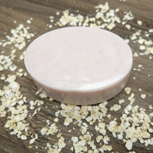 Puede incluir: Una pastilla de jabón ovalada de color rosa pálido, rodeada de copos de avena dispersos, descansa sobre una superficie de madera oscura. El jabón tiene un aspecto liso y ligeramente brillante, lo que sugiere una fórmula hidratante. La imagen destaca productos naturales para el cuidado de la piel.