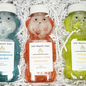 Puede incluir: Tres botellas de baño de burbujas con forma de oso en azul, naranja y verde, cada una con una tapa blanca. Las botellas están etiquetadas como "Asian Sandalwood", "Cashmere Cedar" y "Mahogany Coconut" baño de burbujas. Las botellas están llenas de líquido.