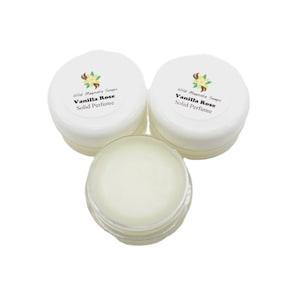 Op de afbeelding: Drie kleine, witte potjes vaste parfum. Twee zijn gesloten, met de tekst "Wild Magnolia Soaps Vanilla Rose Solid Perfume" op de deksels. Het derde potje is open en toont het vaste parfum.