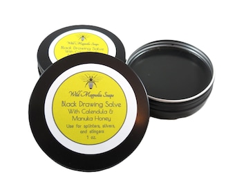 Op de afbeelding: Drie zwarte blikken van Wild Magnolia Soaps Black Drawing Salve. De blikken hebben gele etiketten met de productnaam, ingrediënten en instructies. Het etiket bevat ook een bijengrafiek en geeft aan dat het product 28 gram bevat.