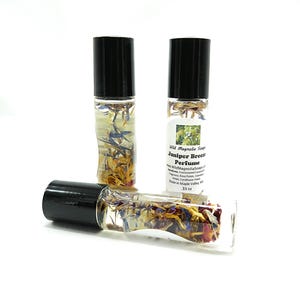 Puede incluir: Tres frascos de perfume roll-on de vidrio transparente con tapas negras. Los frascos contienen flores secas y están etiquetados como "Juniper Breeze Perfume" por Wild Magnolia Soap. Los frascos son de .33 oz.