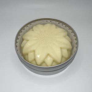 Peut inclure: Une barre de lotion solide jaune clair en forme de tournesol est placée à l'intérieur d'une petite boîte ronde en argent. La barre de lotion est nichée dans une doublure en papier blanc. Le fond est blanc uni.