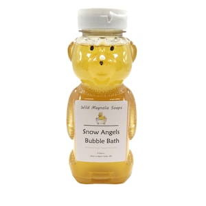 May include: A clear, bear-shaped bottle filled with yellow liquid, topped with a white cap. The label reads "Snow Angels Bubble Bath" from Wild Magnolia Soaps. The bottle contains 8 fluid ounces.