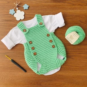Op de afbeelding: Een handgemaakte baby-outfitset, bestaande uit een lichtgroene gehaakte romper met houten knopen, gecombineerd met een wit shirt met lange mouwen. Een bol groen garen en een haaknaald zijn ook aanwezig.